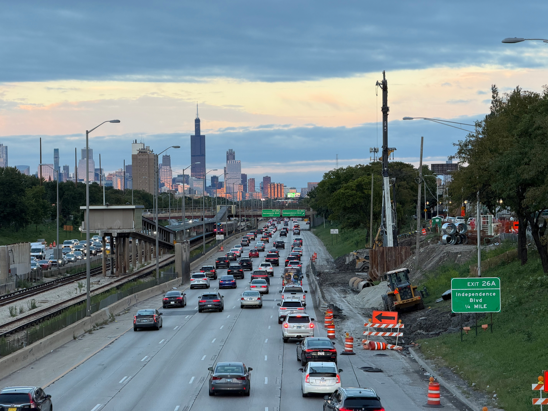 A busy Eisenhower expressway in an urban area shows heavy traffic with construction work taking place on the right side, and a Chicago skyline visible in the background.