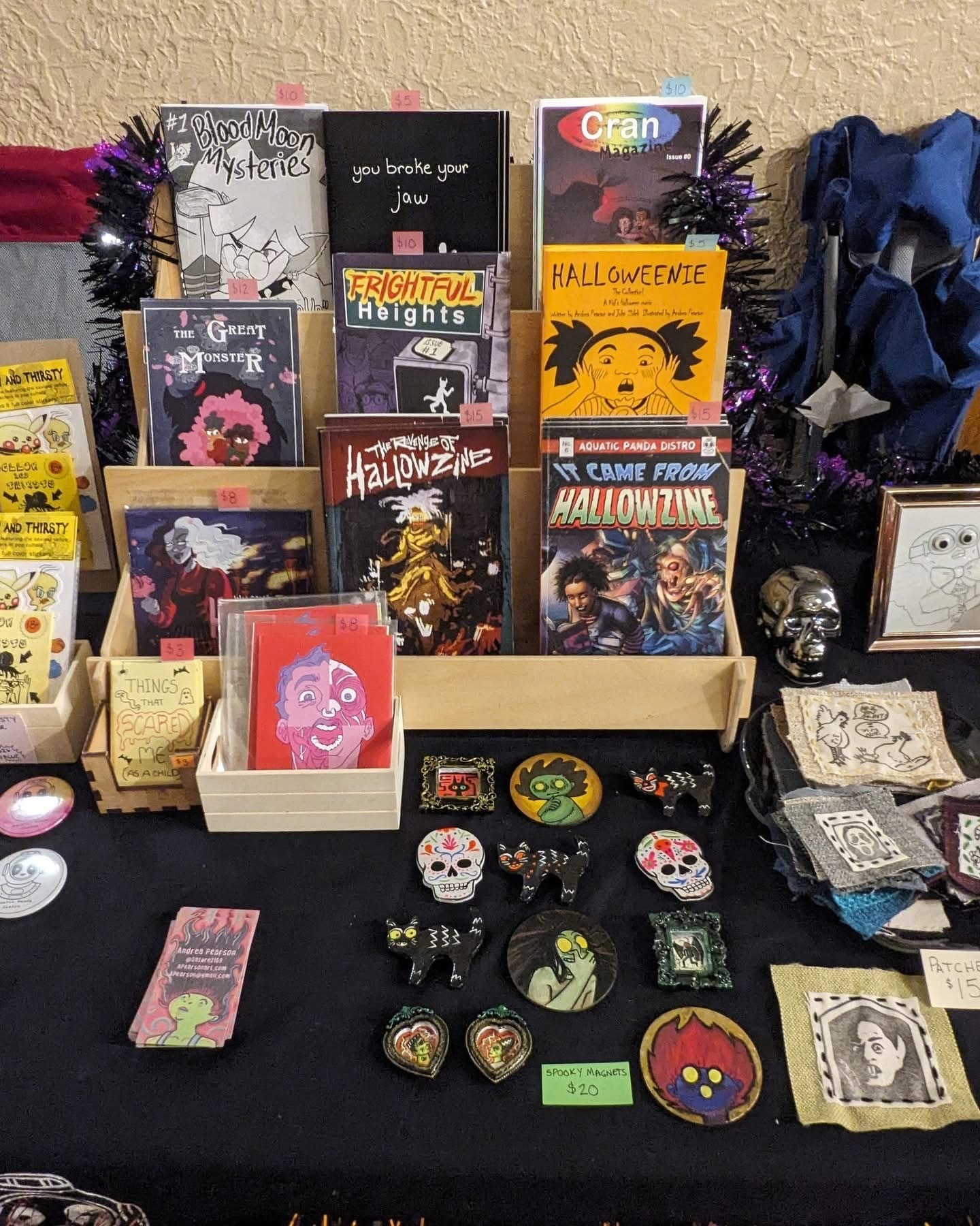 A table displays various spooky and Halloween-themed zines, pins, patches, and small art pieces arranged neatly for sale.