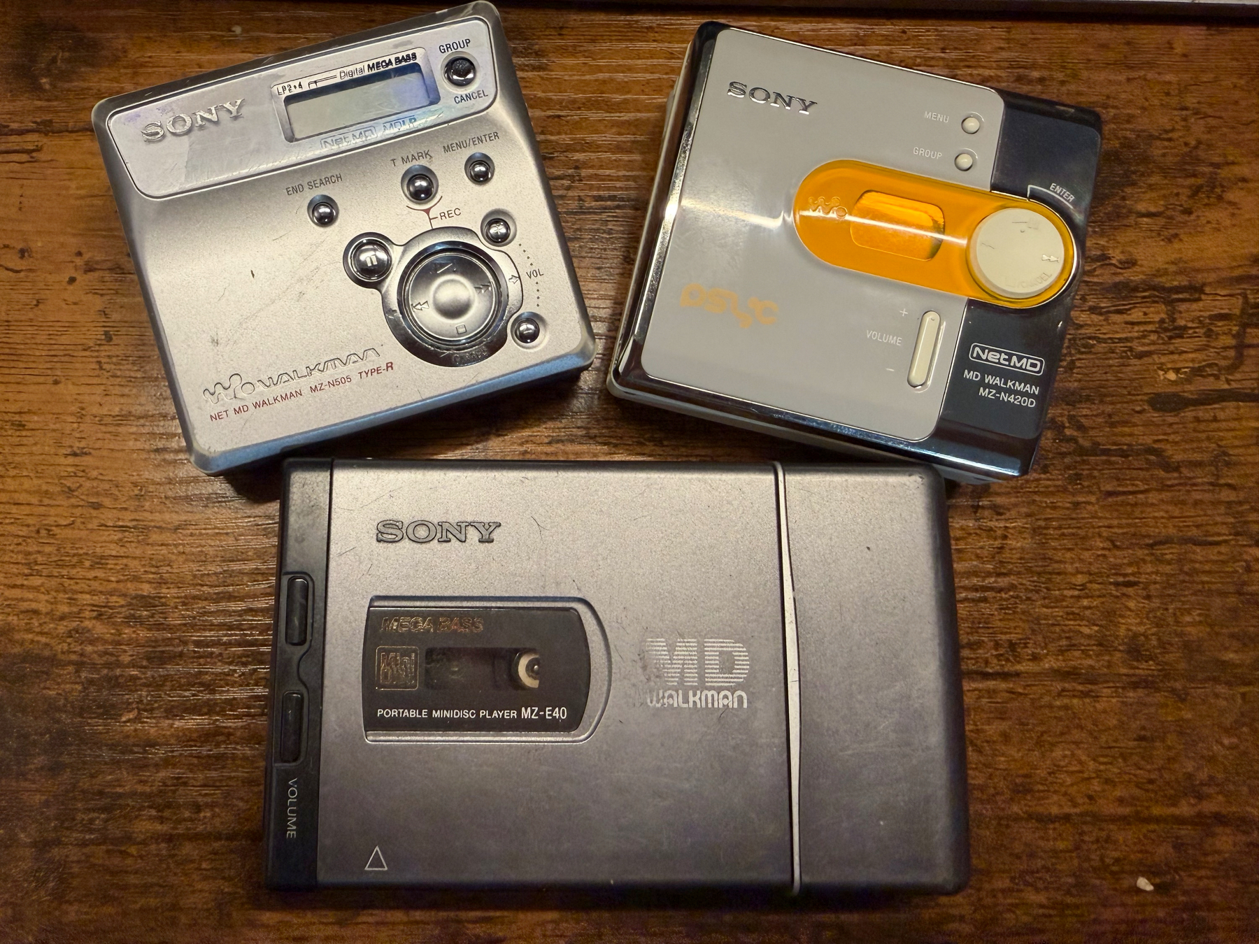 Three vintage Sony Walkman minidisc portable music players are displayed on a wooden surface. As sony MZ-N505 top left, a sony MZ-N4200 nest to it and both are above a classic Somy MZ-E40.