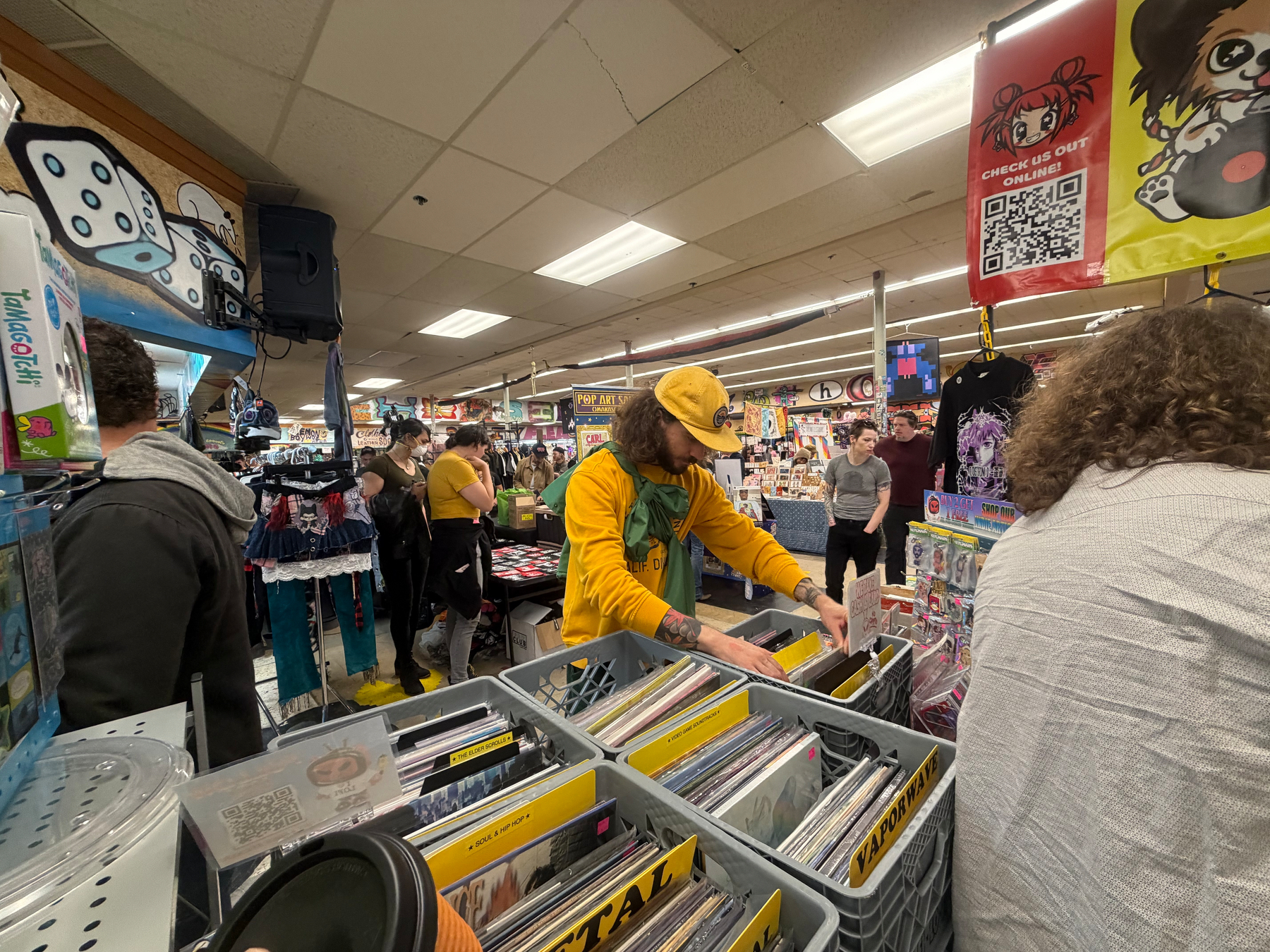 A person wearing a yellow hat and jacket is browsing through vinyl records in a busy store like space filled with various merchandise and other shoppers.