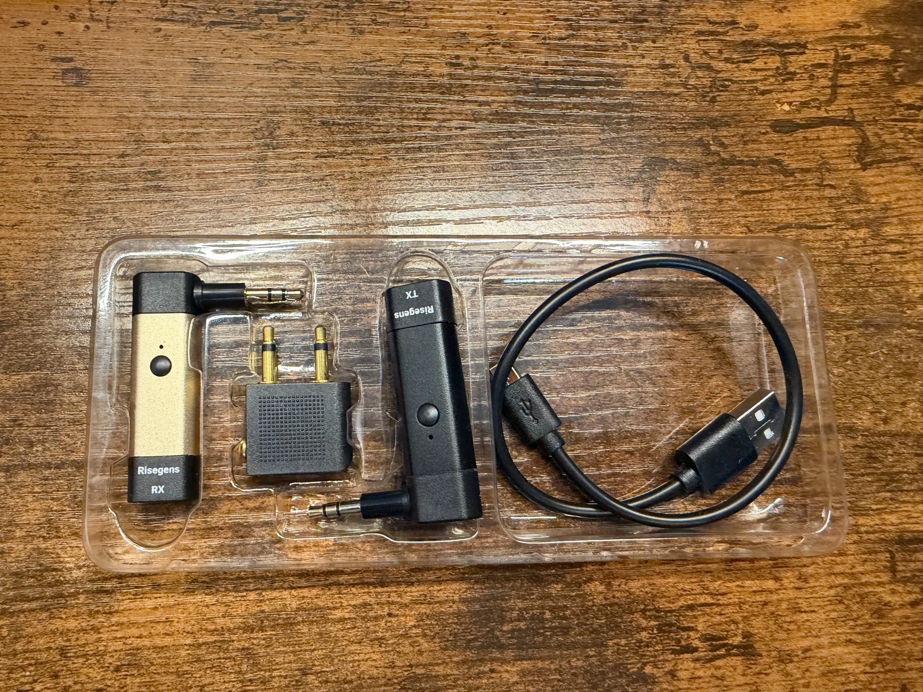 A set of Risegens bluetooth accessories including two adapters labeled RX and TX, a dual plug airplane adapter, and a USB charging cable are arranged on a wooden surface.