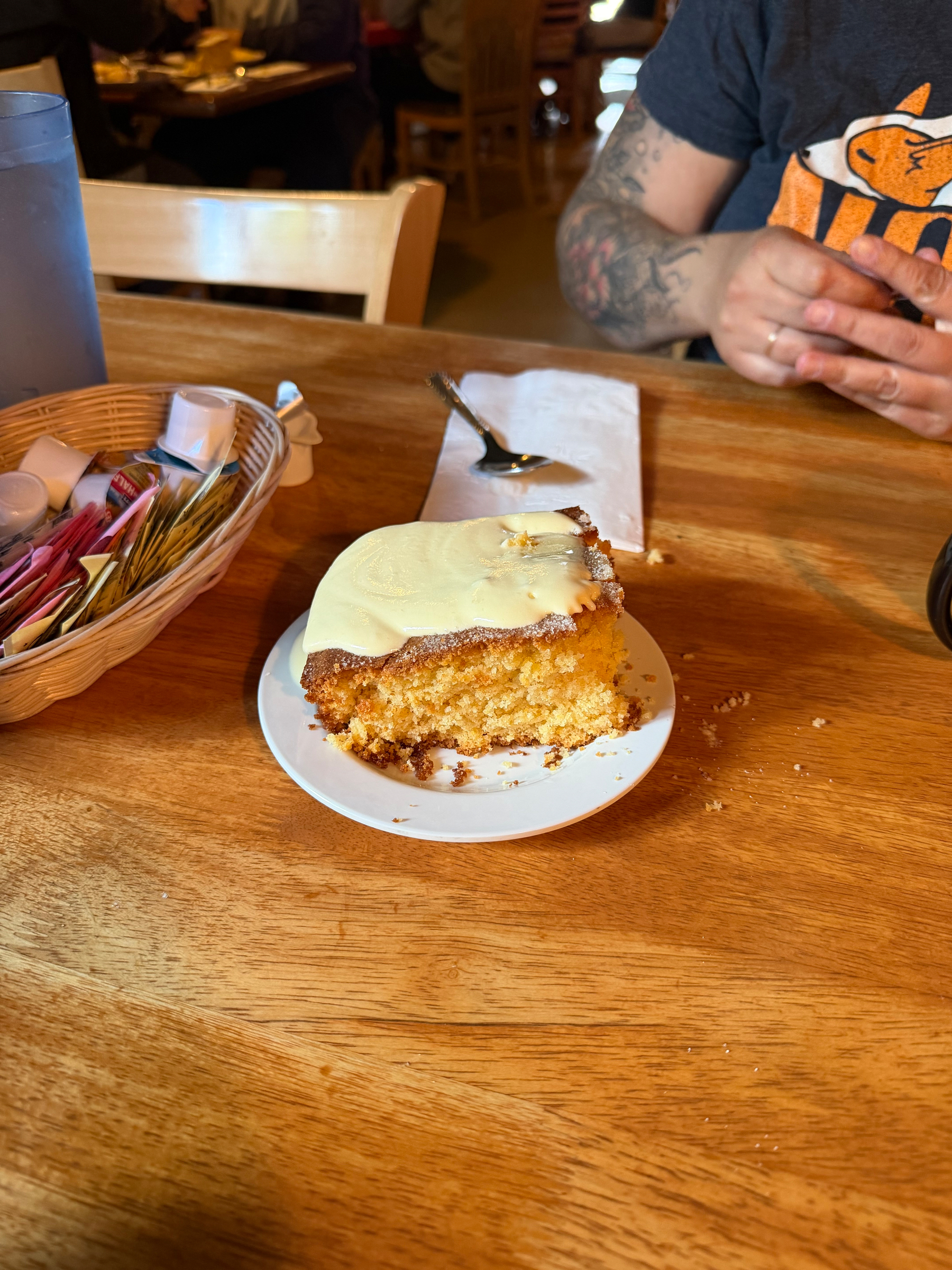 A slice of gigantic corn cake, topped with butter, sits on a white plate atop a wooden table, near a basket of condiments and Ami’s arm is in the background.