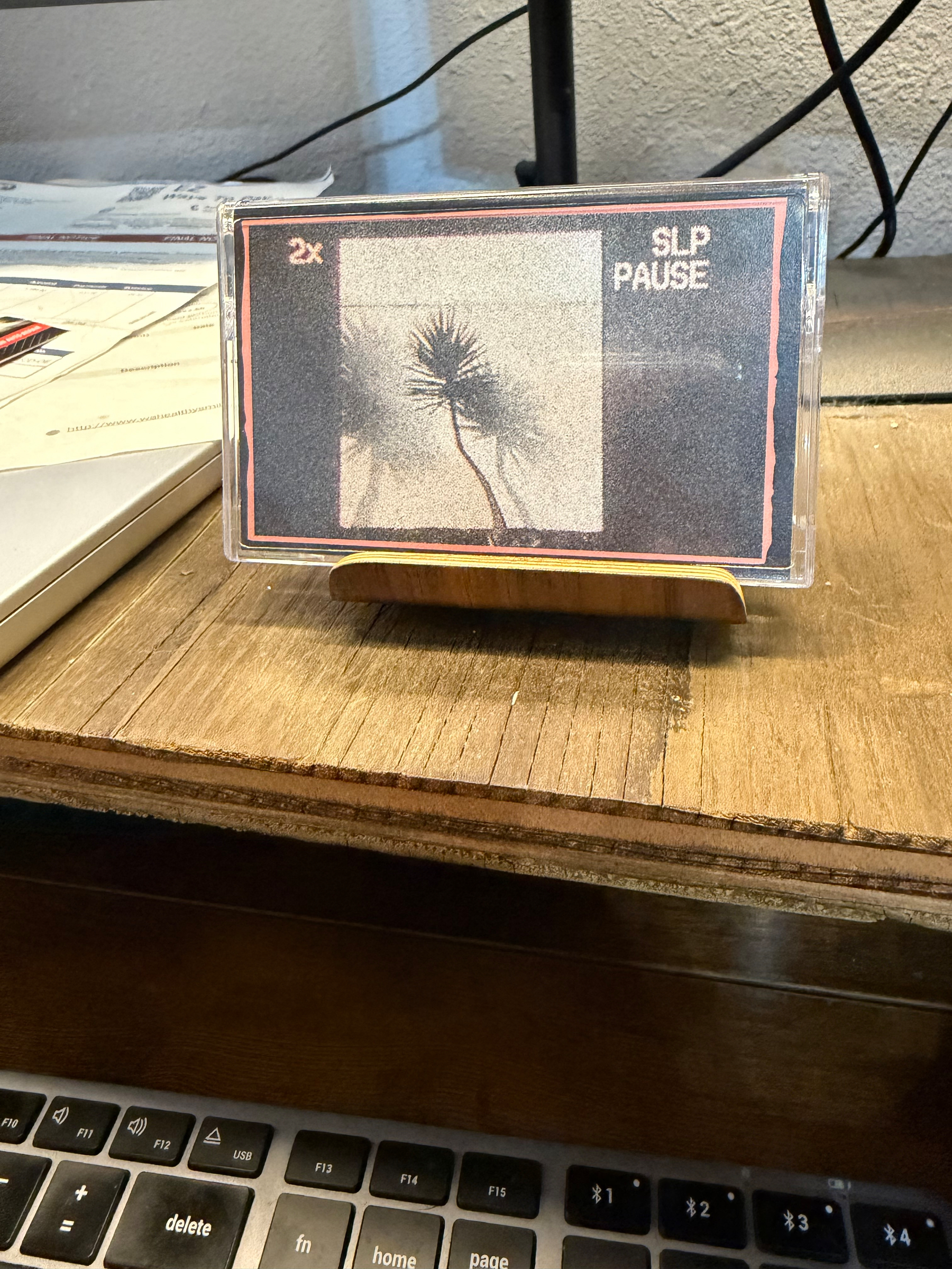 A cassette tape case displaying minimalist artwork with a silhouetted plant on a wooden stand near a keyboard.