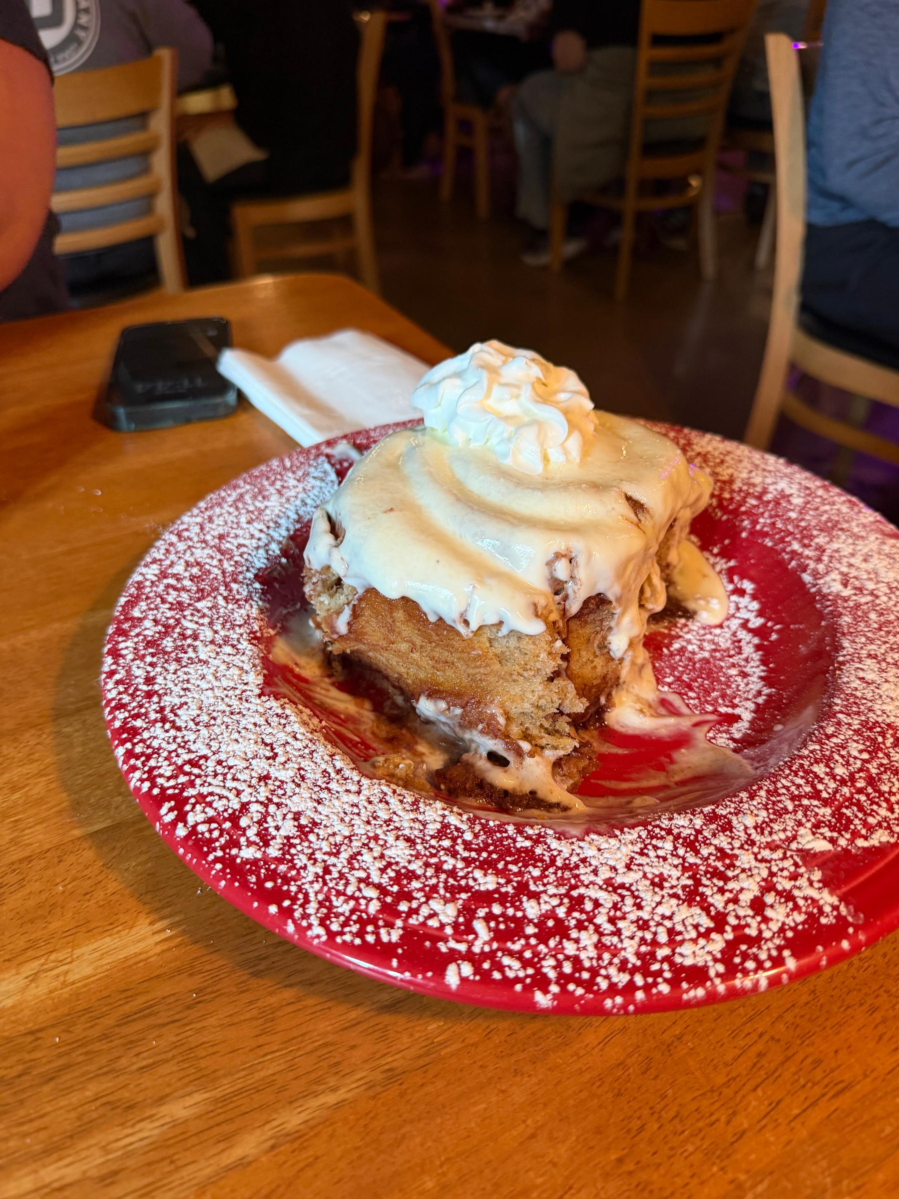 A cinnamon roll topped with cream cheese icing and whipped cream is served on a red plate dusted with powdered sugar.