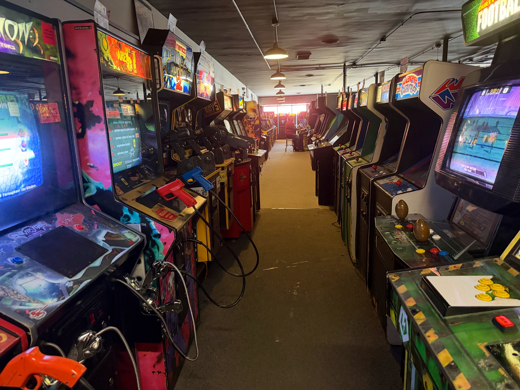 A long aisle is lined with vintage arcade game machines on both sides in a dimly lit room.