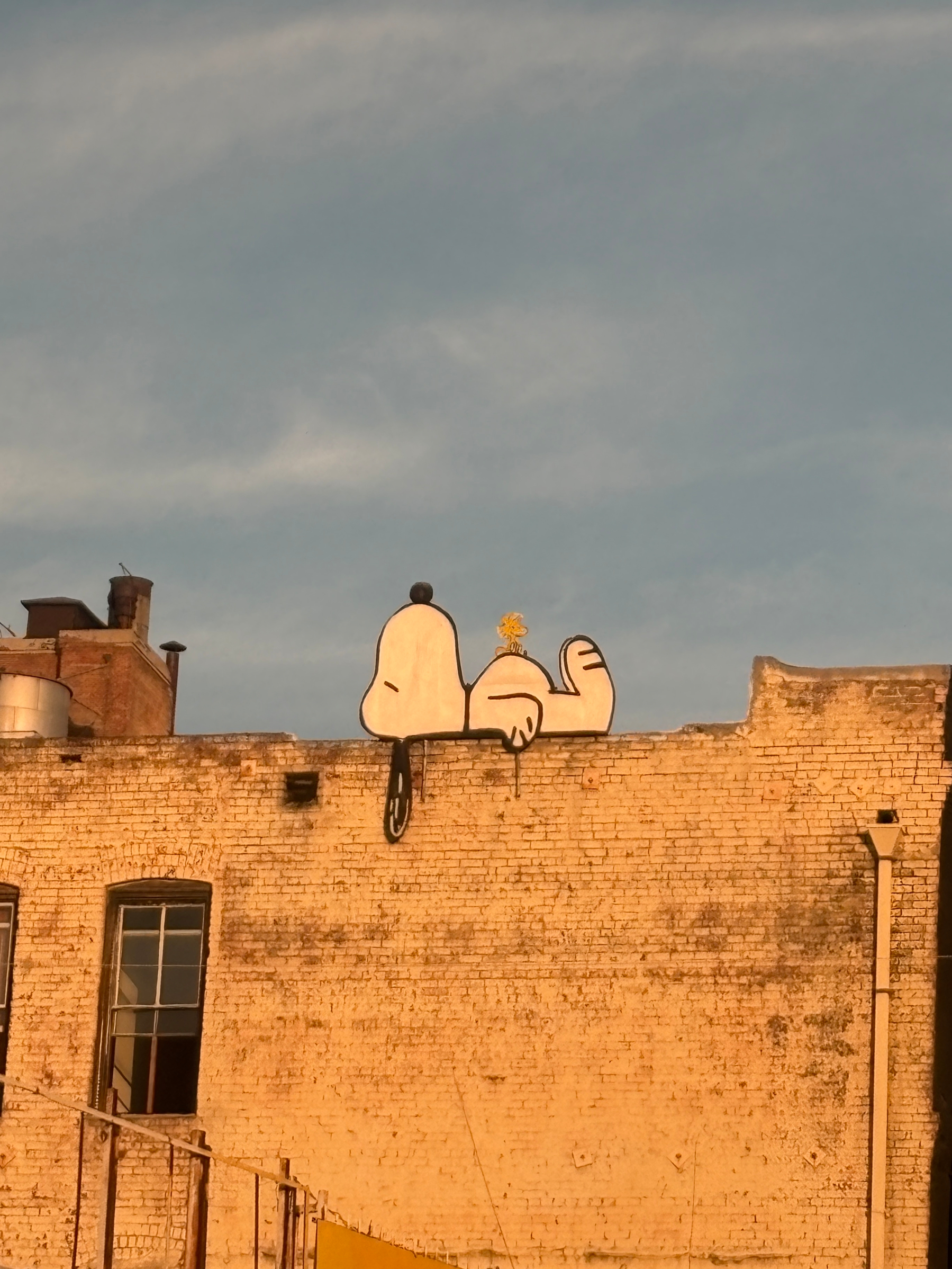 A Snoopy sign  lying on his back with Woodstock on his belly is painted on the top edge of a brick building. 