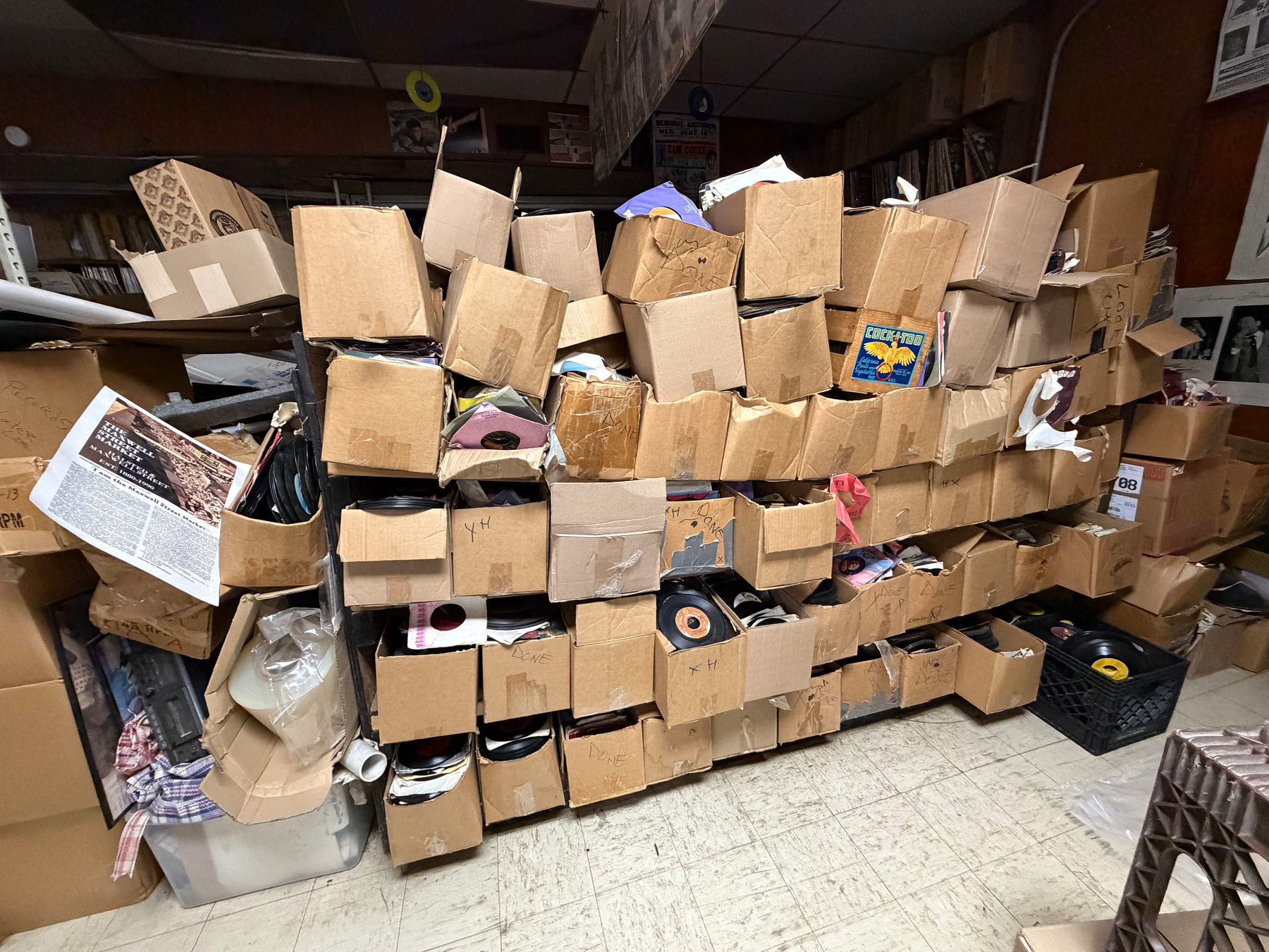 A large collection of cardboard boxes filled with vinyl records is stacked against a wall inside a cluttered room.