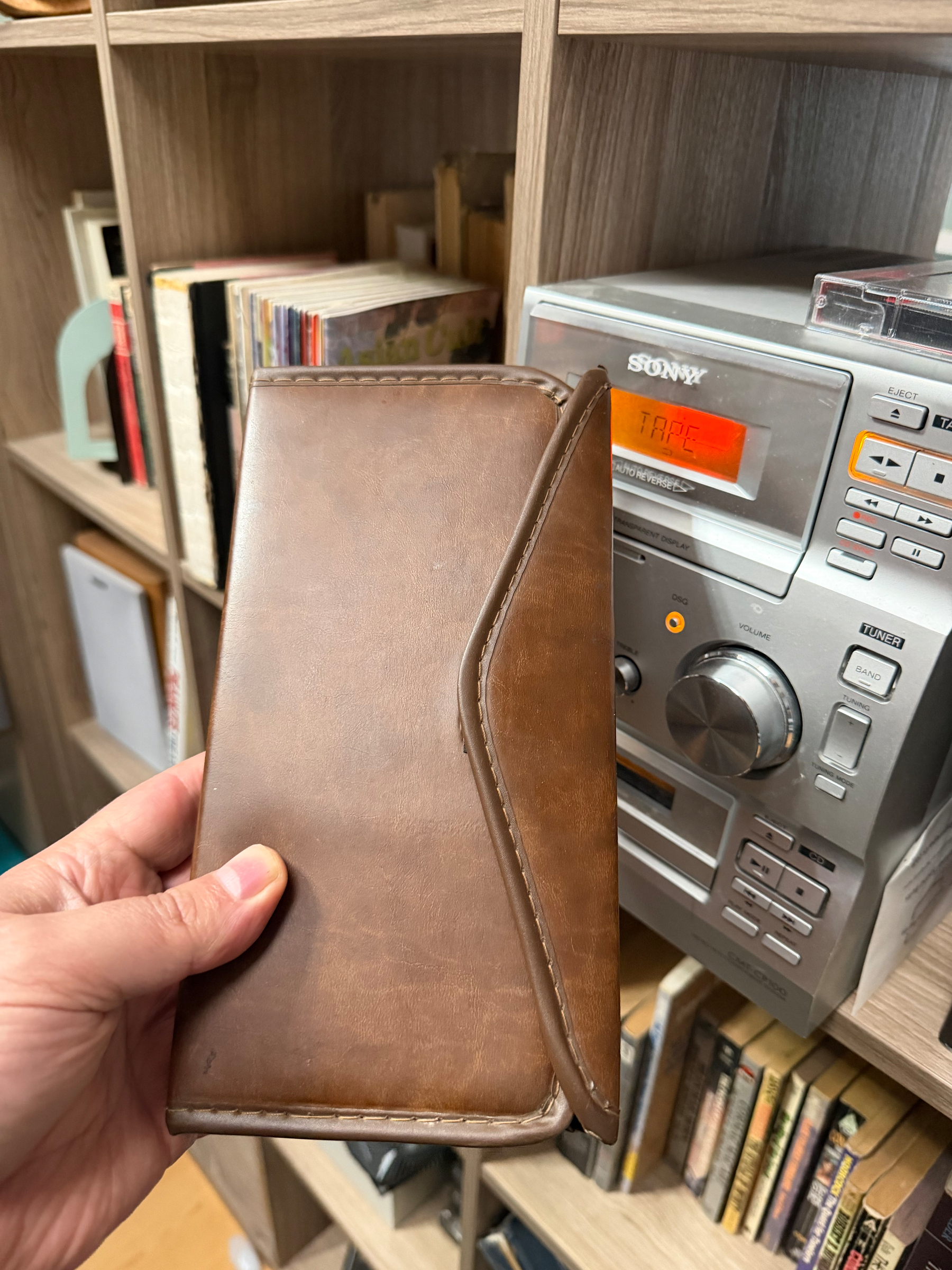 A brown leather tape case is being held in front of a shelf with books and a stereo system.
