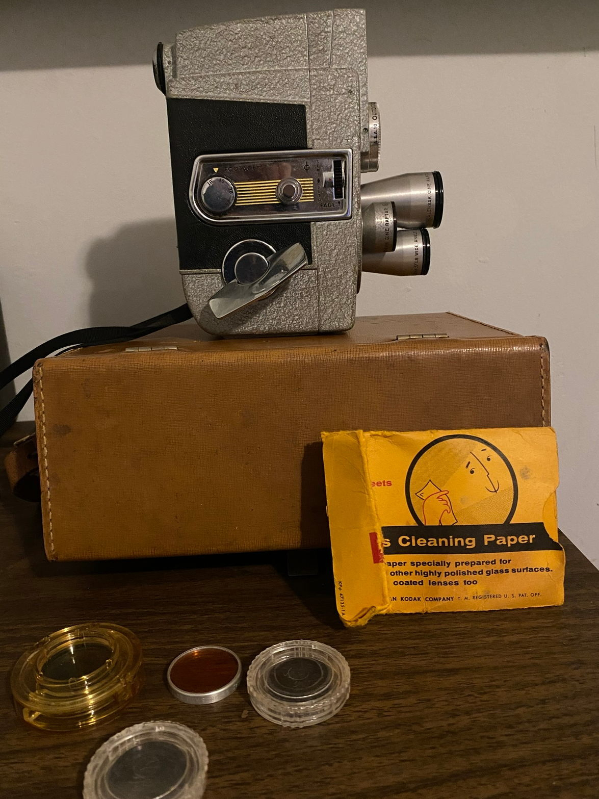 A vintage film camera is placed on a brown case, accompanied by various camera accessories and a yellow Kodak cleaning paper packet.