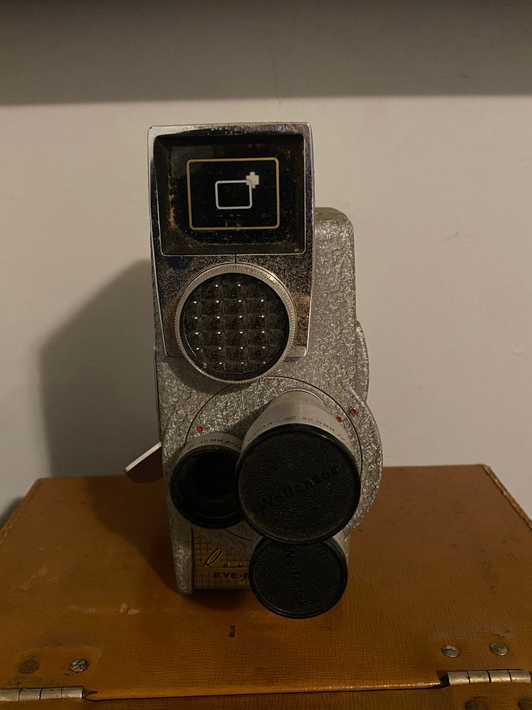 A vintage movie camera with multiple lenses and a weathered exterior is placed on a brown surface against a plain wall.