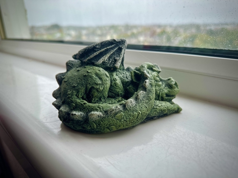 Model of a sleeping, green, dragon, on a window sill. Evidence of rain on the window pane.