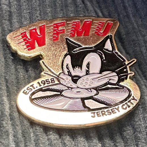 Metal WFMU badge featuring a cartoon cat biting a record.