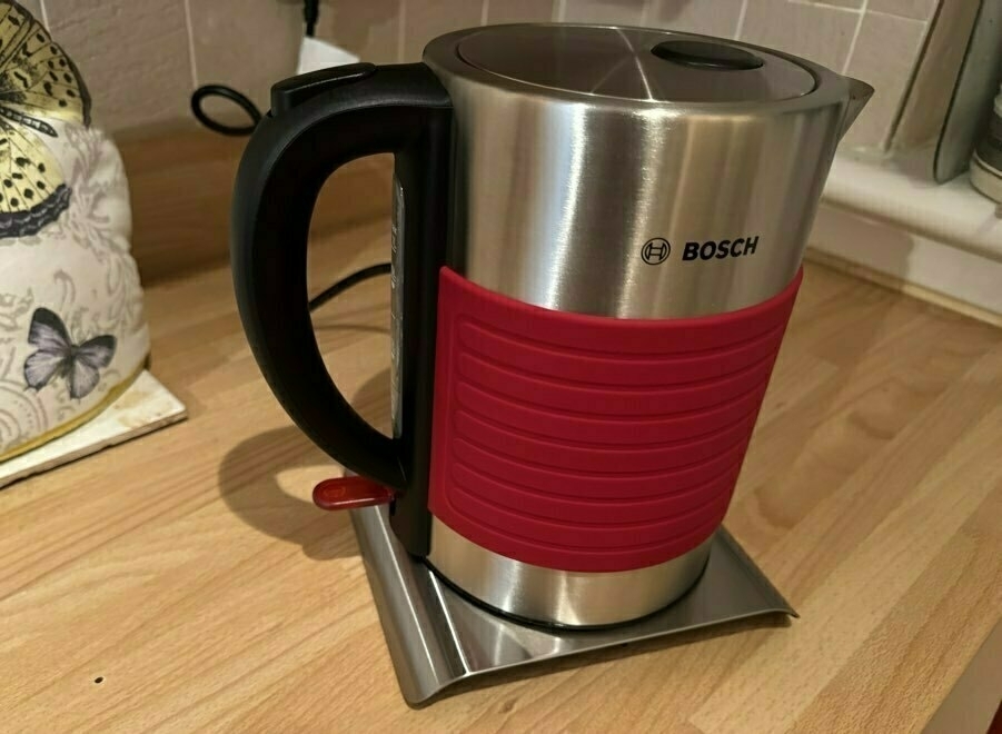 A stainless steel electric kettle with a red central band is placed on a countertop.