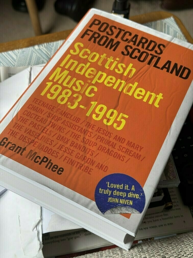 A book titled Postcards from Scotland: Scottish Independent Music 1983-1995 by Grant McPhee is seen, placed atop some papers.