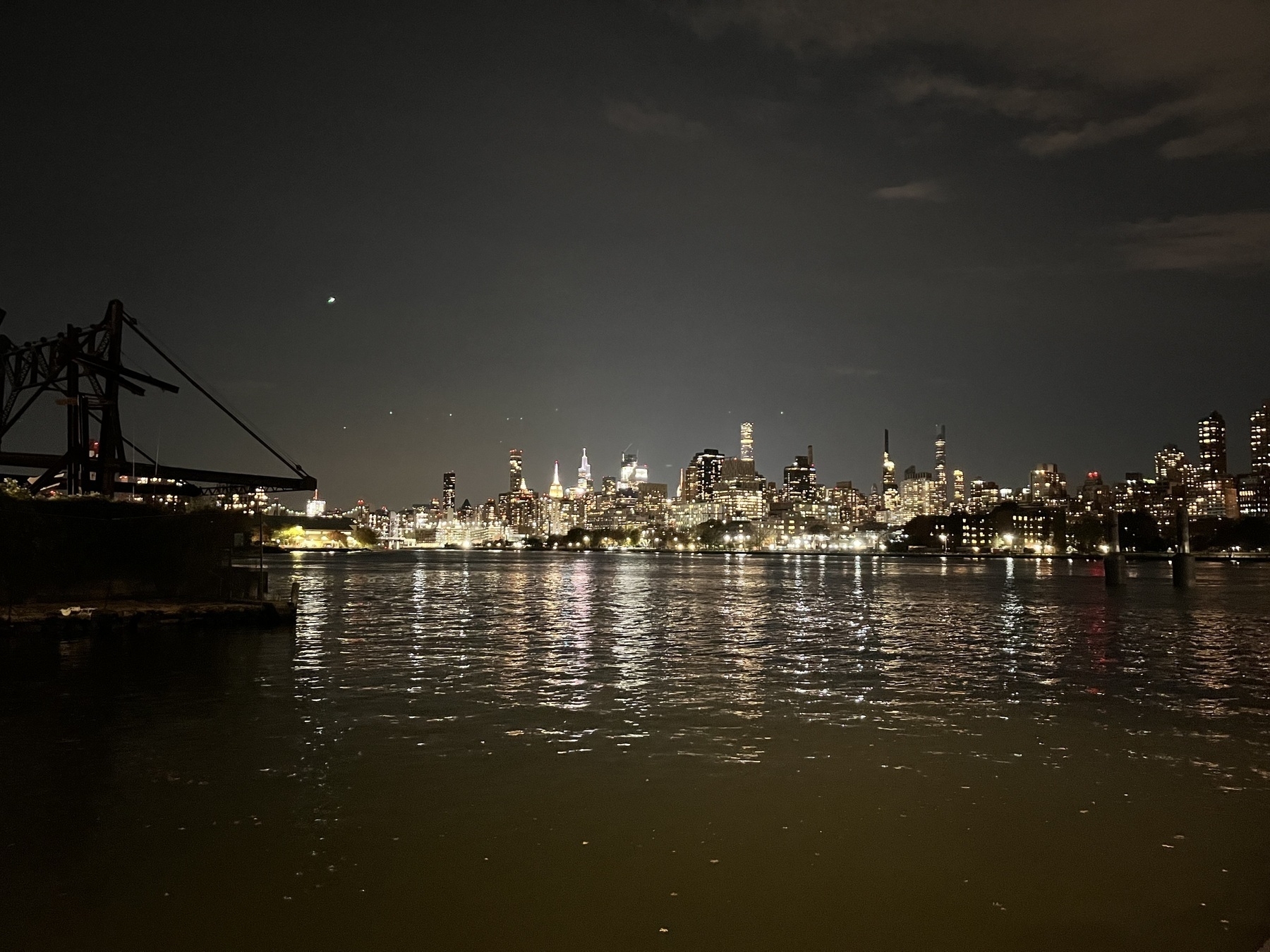 New York City skyline at night, sepia almost, with rippling water in the foreground and the shadow of a raised dock apparatus. 