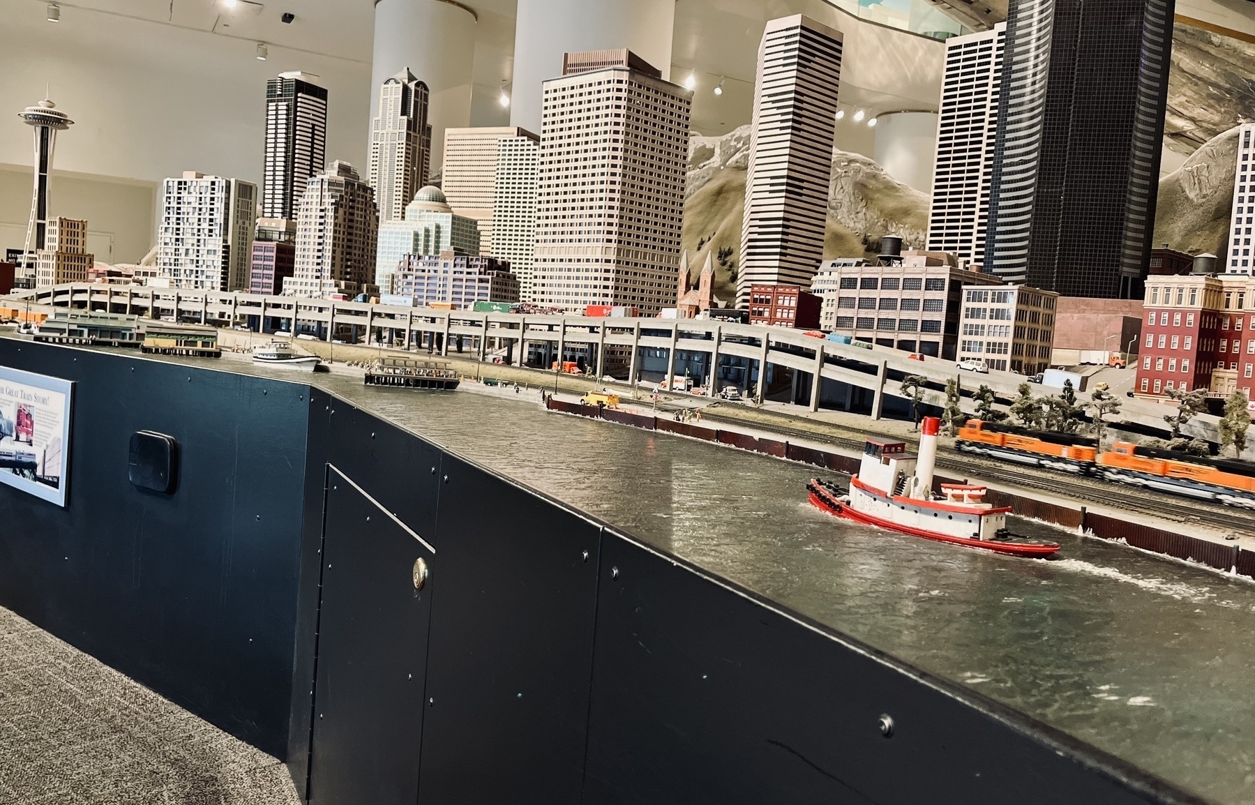 A diorama of Seattle. Builds in the background, river with a boat in the foreground, train in the middle. You can see it’s a small scale model in a large room. 