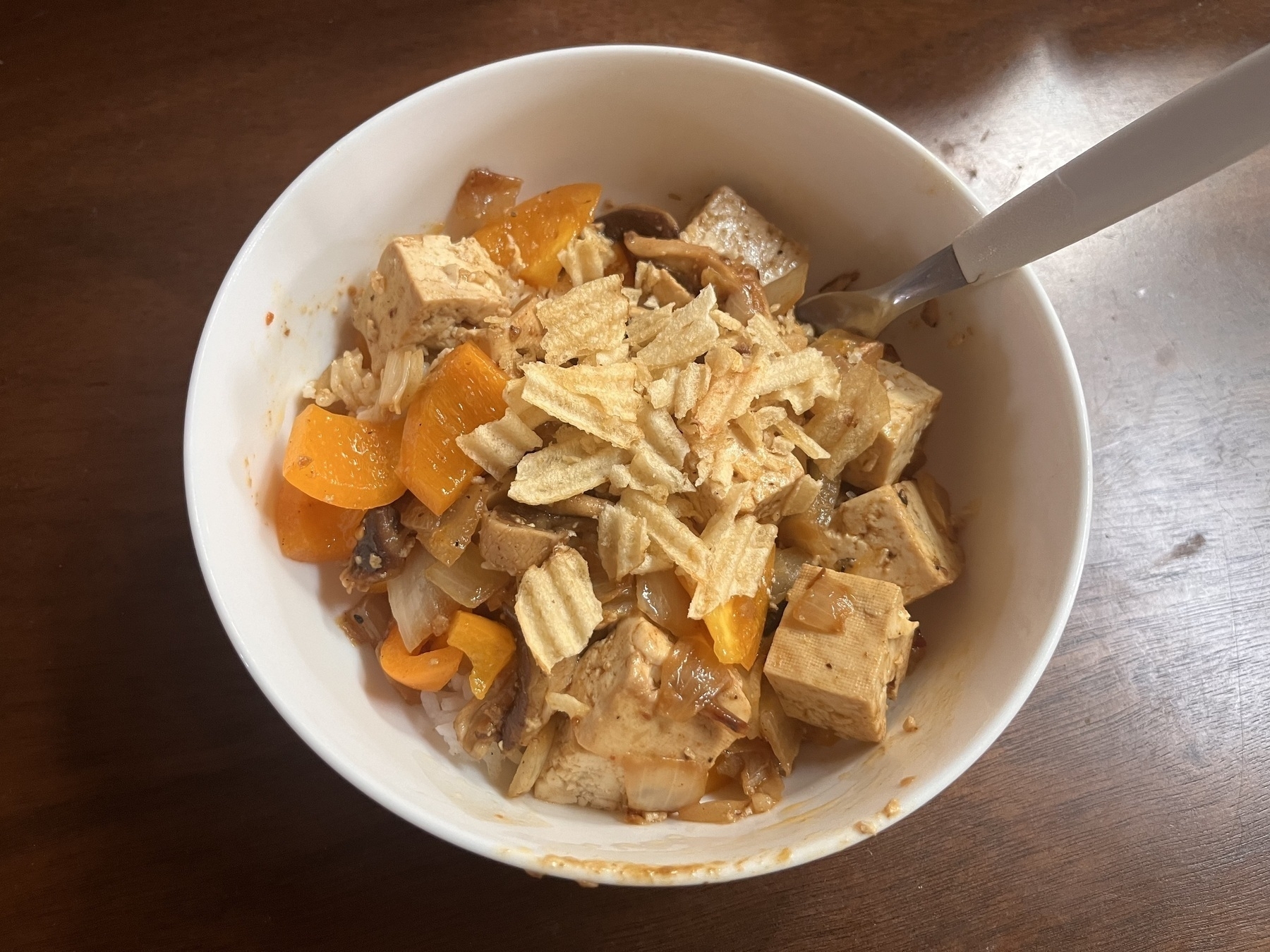 A bowl of rice with stir fried orange peppers, tofu and mushrooms and crushed potato chips on top. 