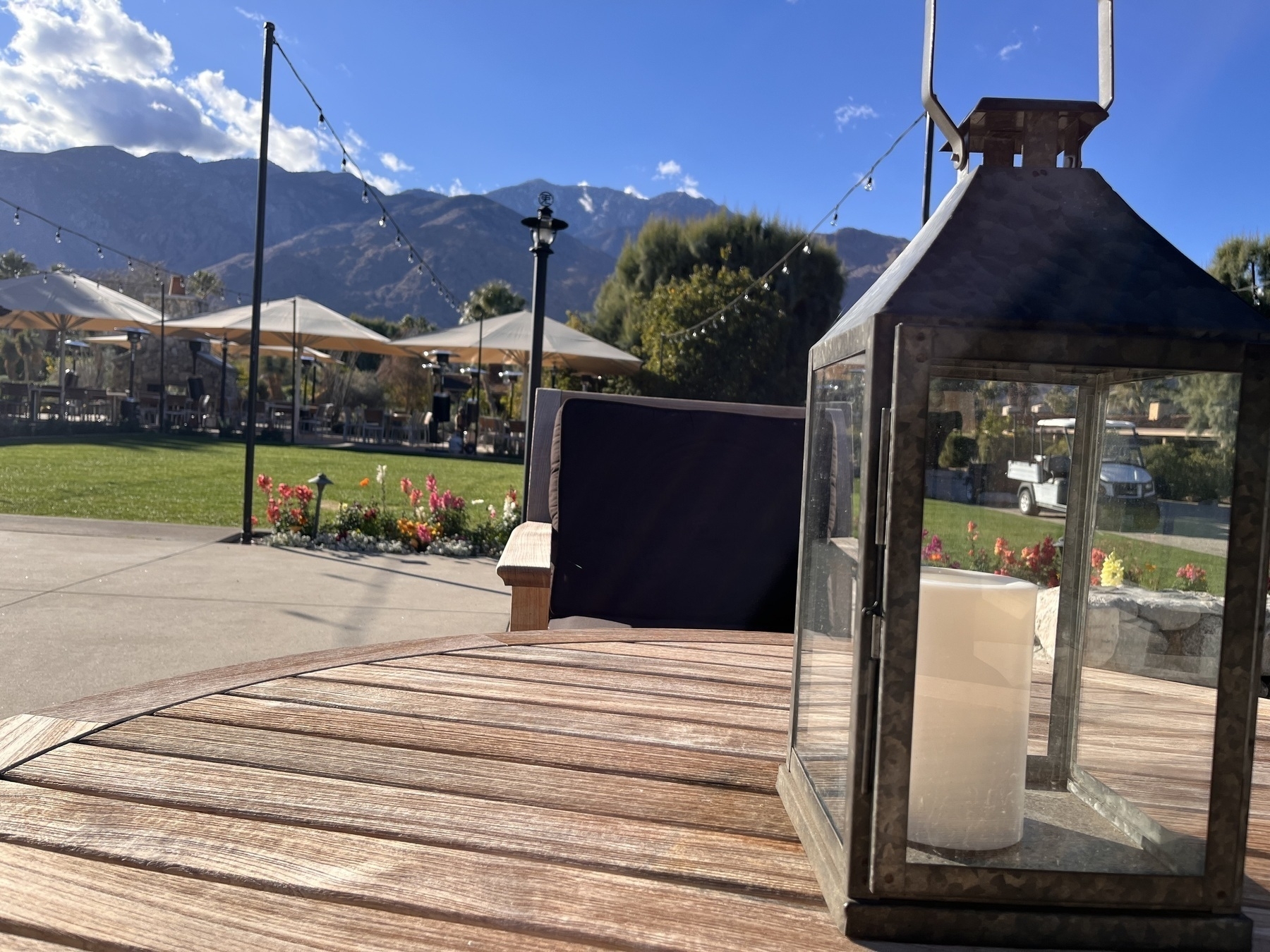 A lamp in the foreground on a table with a green lawn and flower beds and peaked buildings in the middle distance. Mountains rise in the background against a bright blue sky. 