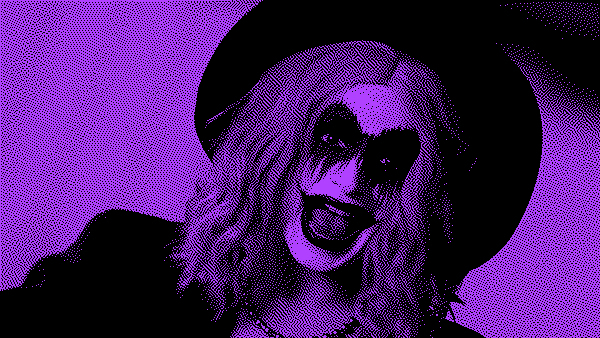 Grainy black and purple photo of the people&#39;s joker, the titular character made up in joker drag, smiling in a wide brimmed hat.