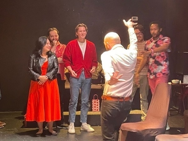 A photo of me on stage in a black box theater, shot from behind. I'm a bald, aging white man. I'm wearing black jeans and a pastel pink dress shirt. My hands a splayed out in eggagerated gestures. Some of my improv teammates, an Asian woman, three white men, and a black man, look on.