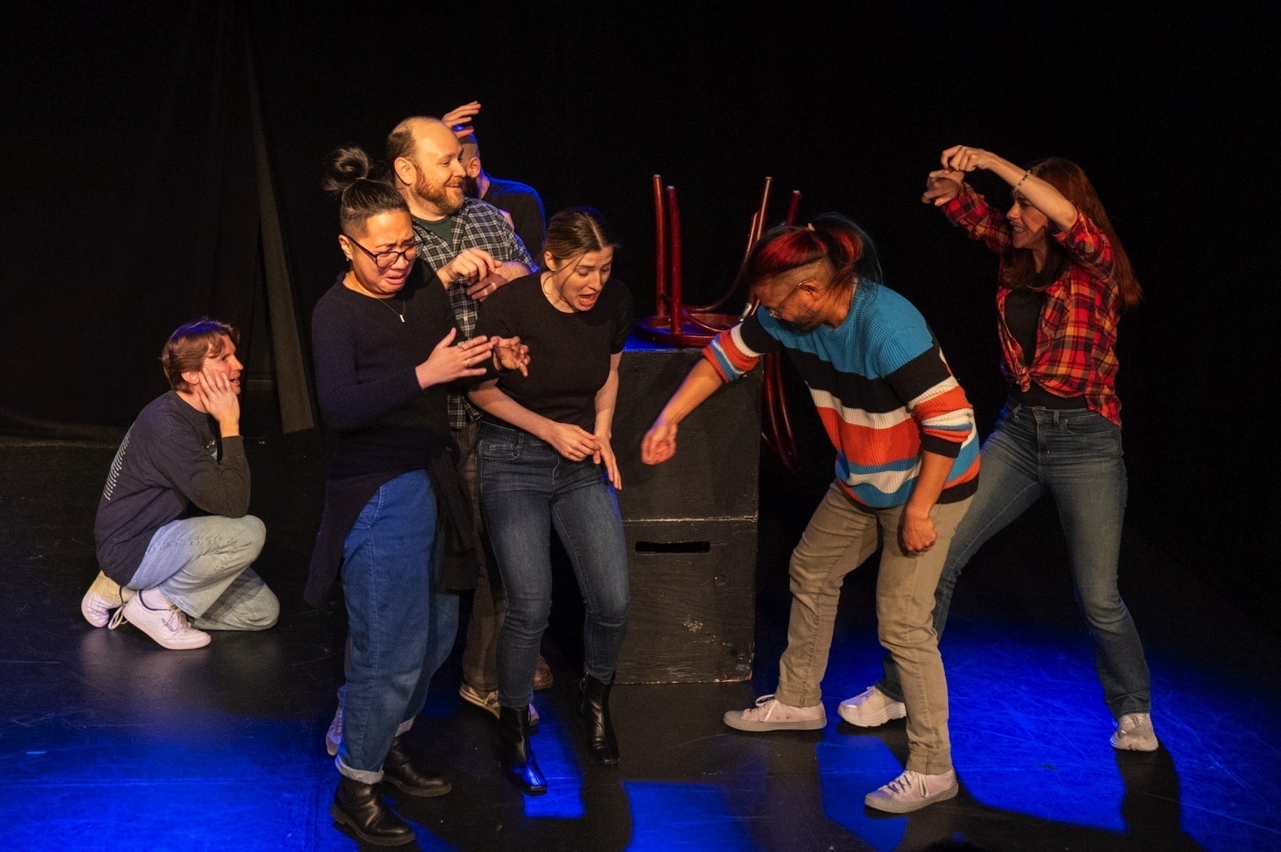 A group of six people engaged in an animated theatrical performance on a stage. Some individuals are crouched or bent over, expressing mock horror or surprise, while others stand with exaggerated gestures of fear or caution. They appear to be reacting to an unseen object or event on the right side of the stage. The stage has a plain black background and minimal props, with a single black box at the center. The actors are dressed in casual clothing and the lighting casts a blue hue over the scene.