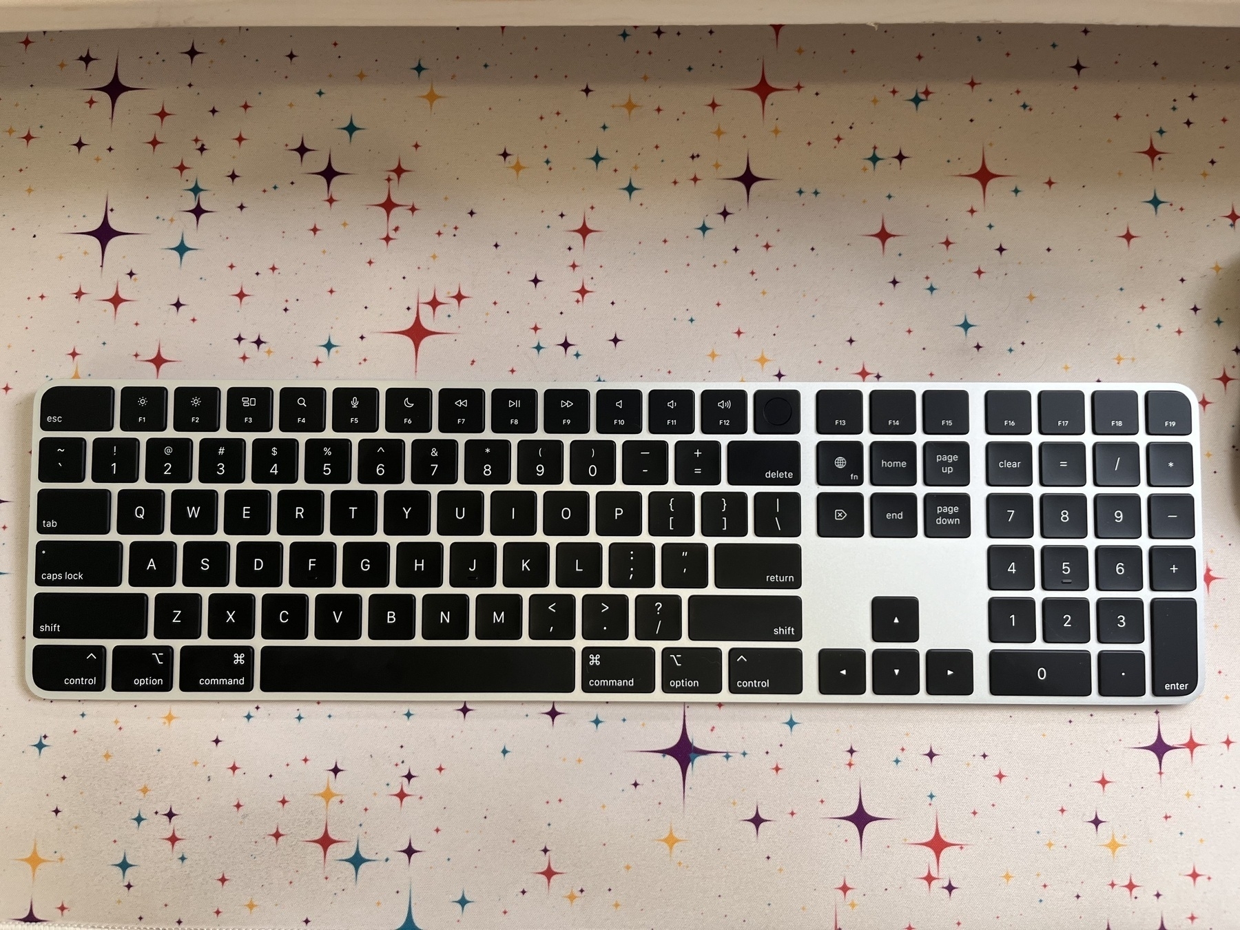 The black apple Magic Keyboard with the full numpad resting on a colorful mat with a star pattern. 