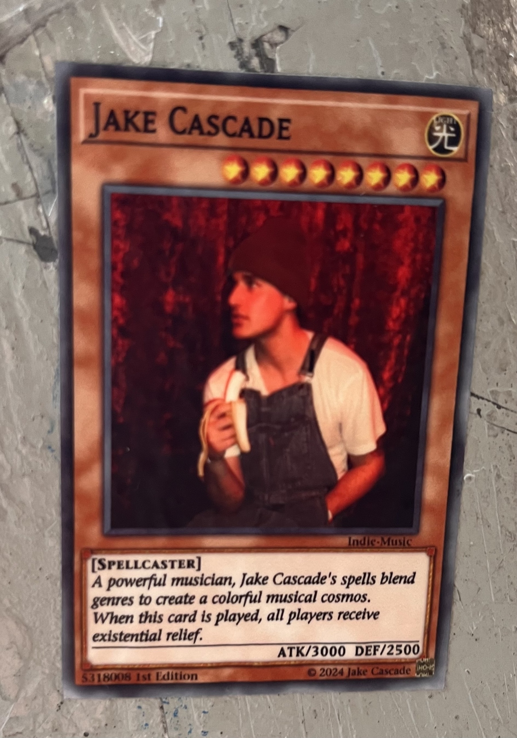 A poster on a light post made to look like a fake magic the gathering card. It’s called Jake cascade and features a picture of a young man wearing overalls and a winter hat and holding a banana. The card text reads: “spellcaster. A powerful musician, Jake cascade’s spells blend genres to create a colorful musical cosmos. When this card is played all players receive existential relief. “