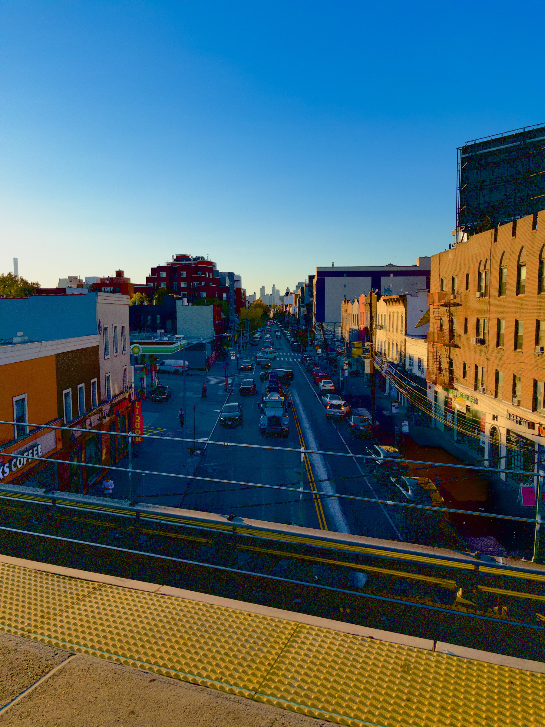 Auto-generated description: A city street view at sunset shows cars driving along a road lined with buildings under a clear blue sky.