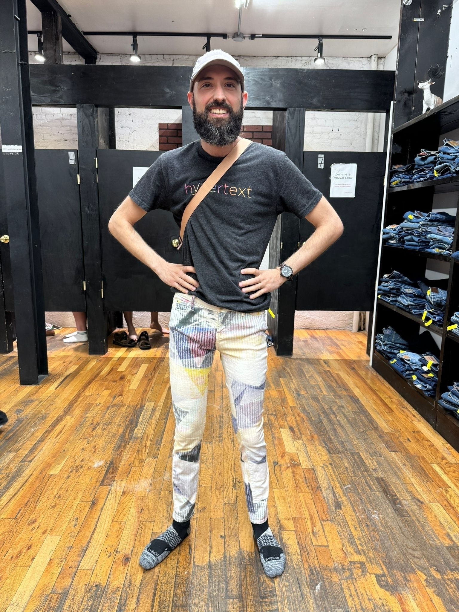 Me, an older white man with a long beard, stands in a thrift shop. My hands are on my hips and I’m smiling. I’m wearing these skin tight white and pastel color splotch pants.