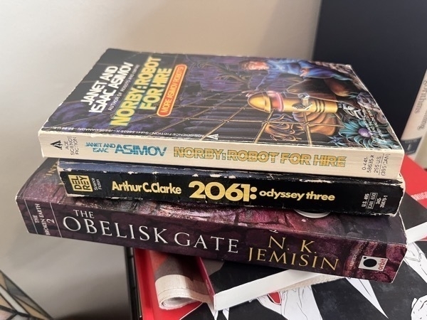 3 books stacked on top of each other: The Obelisk Gate by N.K. Jemisin, 2061: Odyssey Three by Arthur C. Clarke, and Norby: Robot for Hire by Janet and Issac Asimov