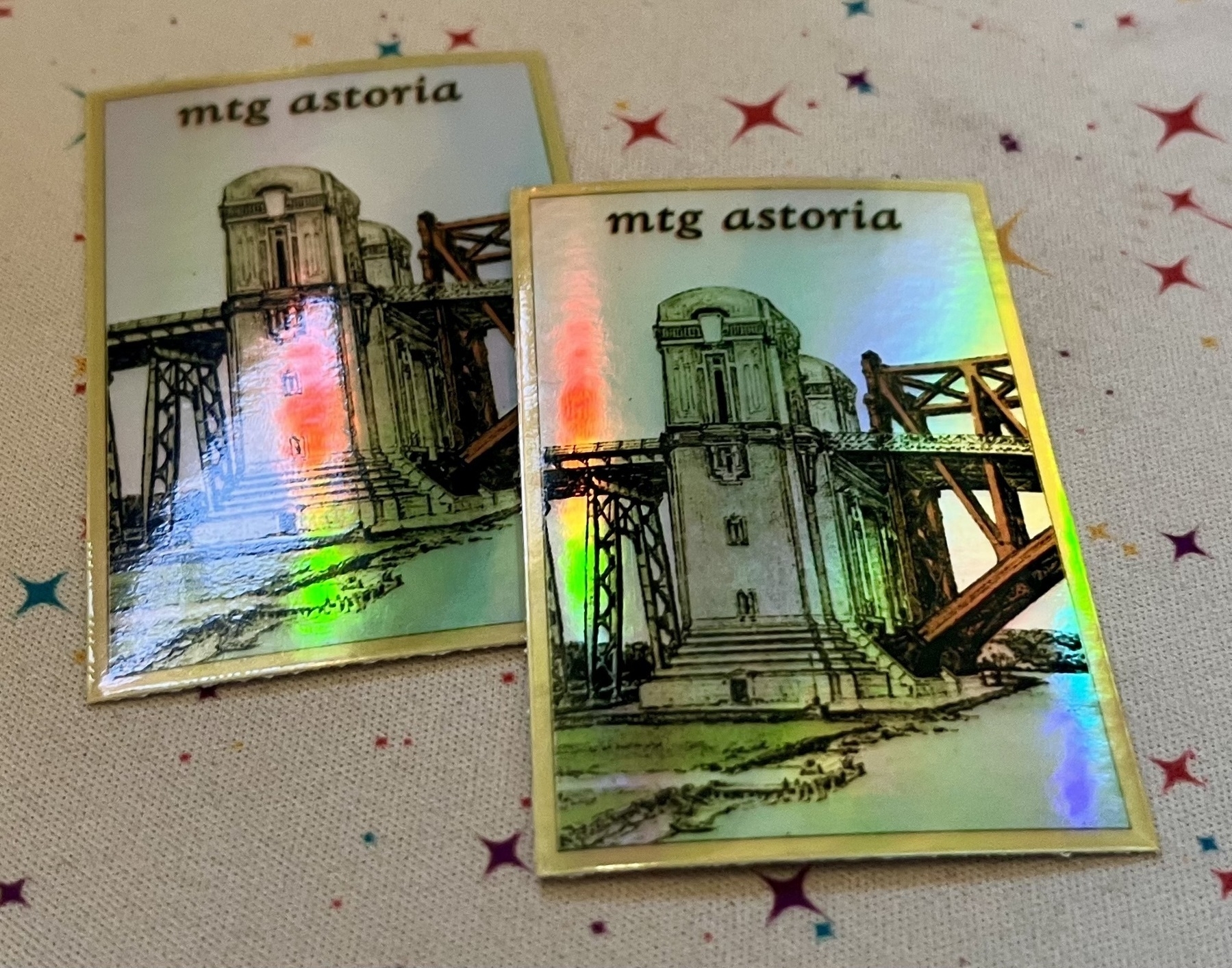 Two foil stickers that say “mtg astoria “ above a drawing of maybe the hellgate bridge. 