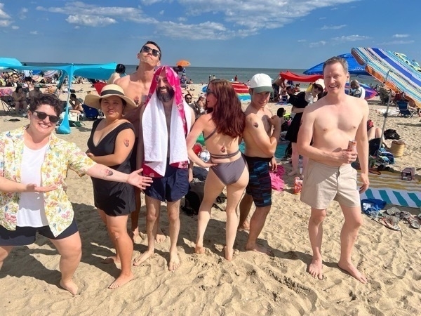 A group of six friends is posing playfully on a crowded beach under a sunny sky. The group includes three men and three women. Each person has a temporary tattoo of Chris' face. One woman on the far left is wearing a floral shirt and shorts, enthusiastically pointing to her left arm. Her left arm has Chris' face near the elblow joint. Next to her is a woman in a black swimsuit and sunhat, posing with her hand on her hip. She has Chris' face on her bicep. In the middle, a man has towel draped over his head. This is Chris. The towel has his face on it. Standing next to him is a woman in a gray bikini with her back to the camera, playfully looking over her shoulder. Chris' face is on her lower back. A man in sunglasses and a baseball cap stands behind them, flexing his arm. On the far right, a shirtless man in beige shorts smiles while holding a drink can. The beach is bustling with people, colorful umbrellas, and towels.