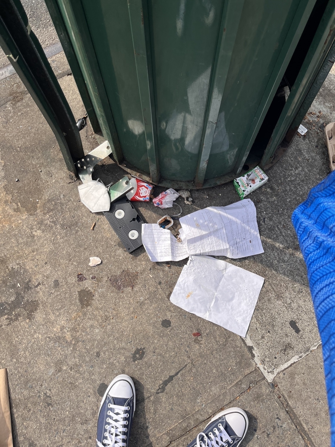 A down shot of a concrete city sidewalk. A garbage can looks in the frame. There’s trash all around it. A grimy VHS lays there amidst the garbage, back up. 