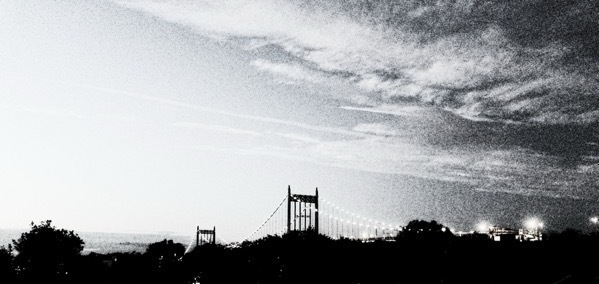 An almost monochrome photo of the RFK bridge in deep shadow. Dramatic clouds streak overhead. There's a ton of film grain.