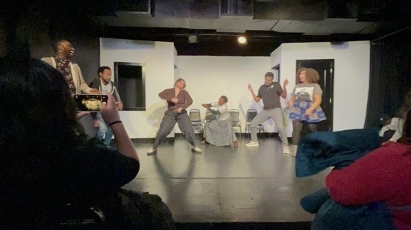 Six black improvisors, half women half men, dancing on a black box theater stage.