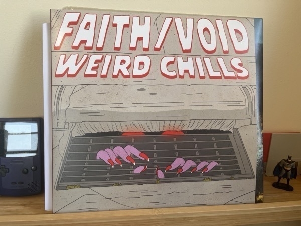 A vinyl record standing against a wall, with a batman and gameboy next to it. The album is called Weird Chills by the band Faith/Void. The cover is a old school horror movie stylized sewer grate, with pink feminine hands with red painted nails coming out of the grate.