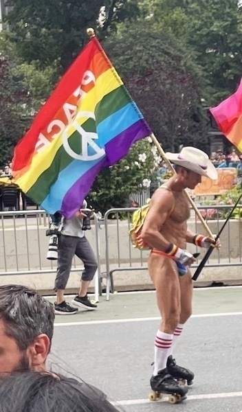 A muscular white man wearing only a thong, a white cowboy hat, and roller skates rolls with a forlorn expression, eyes down. He carries a large peace pride flag.