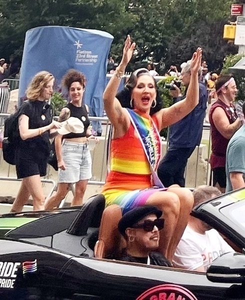 An white woman with primpted black hair wears a bright rainbow form fitting dress. She sits on the back of a car, arms raised mouth open in celebration.