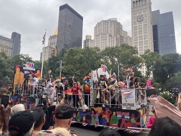 A colorful float is part of a parade, adorned with people waving rainbow flags and holding signs. The float is decorated with vibrant illustrations and a banner reading “NYC Pride 2024.” The participants appear joyful, celebrating and interacting with the crowd. Tall buildings and lush green trees are visible in the background, under a cloudy sky. The scene captures the lively and inclusive spirit of the event.