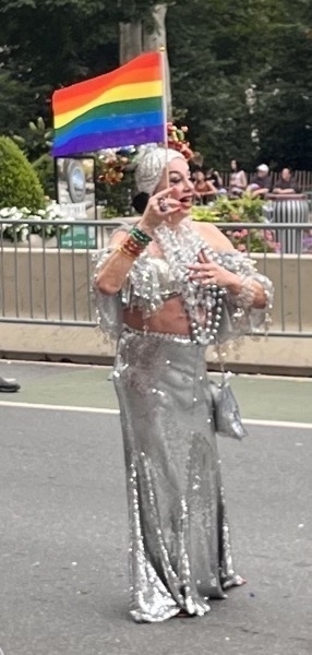 A white drag queen in a fancy silver dress with big silver necklaces, standing tall and holding a pride flag.