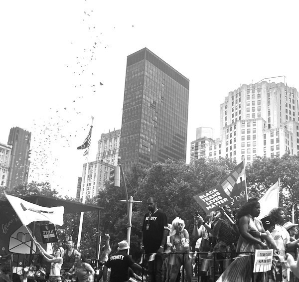 A black and white image of a pride float, many celebrating bodies. The new york city skyline stretches high above the revelers against a pure white sky. A spiral of confetti arcs up from the back of the float, framed in high contrast against the white sky. A Black trans lives matter flag is visible.