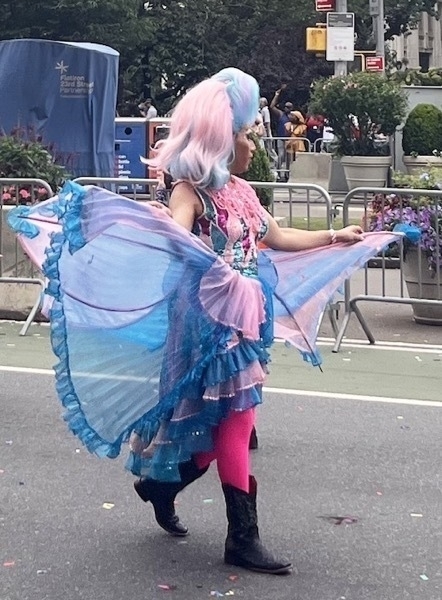 A brown drag queen wears pink and blue fairy wings and corset, with matching hair and big black boots. She walks with purpose.