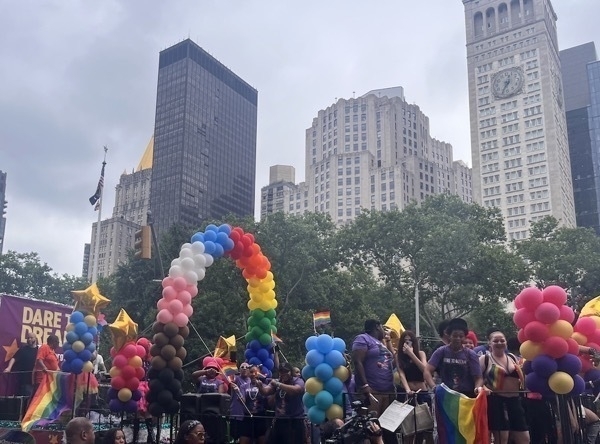 A colorful float with an arc of rainbow colored balloons amidst LGBTQ+ mutli-ethnic celebrants.