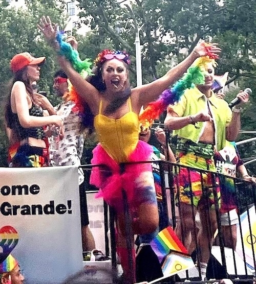 A group of individuals with obscured faces is standing behind a metal barrier at an outdoor event, possibly a parade. They are wearing colorful attire, including rainbow-colored accessories such as wigs, tutus, and flags. One individual in the center wears a yellow top and pink shorts while holding up their hands mid-cheer. A sign with the text ‘Home Grande!’ is visible in the lower left corner, accompanied by rainbow motifs suggesting the event is LGBTQ+ related.