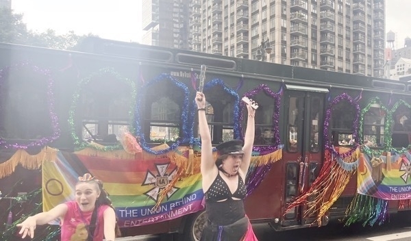 A decorated bus with rainbow colors and the words ‘The Unforgotten’ printed on it is featured in the image. The bus has purple, green, and gold garlands around the edges, and two individuals with obscured faces are standing in front of it. One individual is wearing a black top with a purple skirt and holding up a peace sign, while the other is wearing a black outfit with white polka dots and waving. The background shows high-rise buildings under an overcast sky. The scene appears festive, likely from a parade or celebration, related to LGBTQ+ pride.