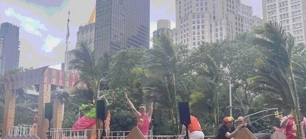 A pride float with palm fronds and pyramids. A man with sunglasses and a pink shirt arms raised yelling with joy is prominantly featured.