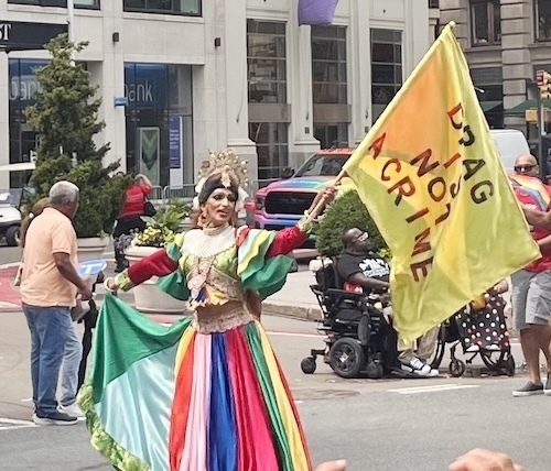 A person in an elaborate rainbow colored dress and makeup waves a flag reading 'drag is not a crime'