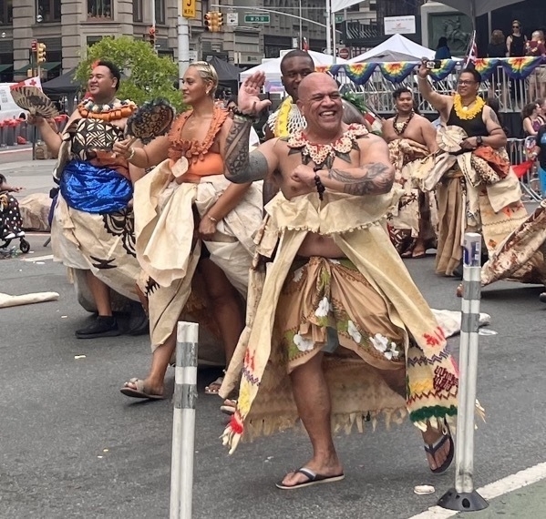 A bald brown man in traditional Somoan tattoos and garb waves with a big smile amidst other similarly dressed celebrants.