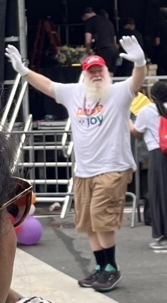 An older white man with a long white beard and a red baseball cap and white gloves and cargo shorts and a pride and joy t-shirt waves, smiling.