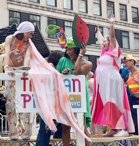 People on a parade float are dressed in colorful costumes. One person on the left is wearing a flowing outfit with a headscarf, while another person in the middle is dressed in green with a large hat adorned with flowers and holding a watermelon prop. A third person on the right is wearing a pink and white dress and has their arms raised in celebration. The float has a sign that reads “Taiwan Pride in NYC.” The background shows a multi-story building with many windows.